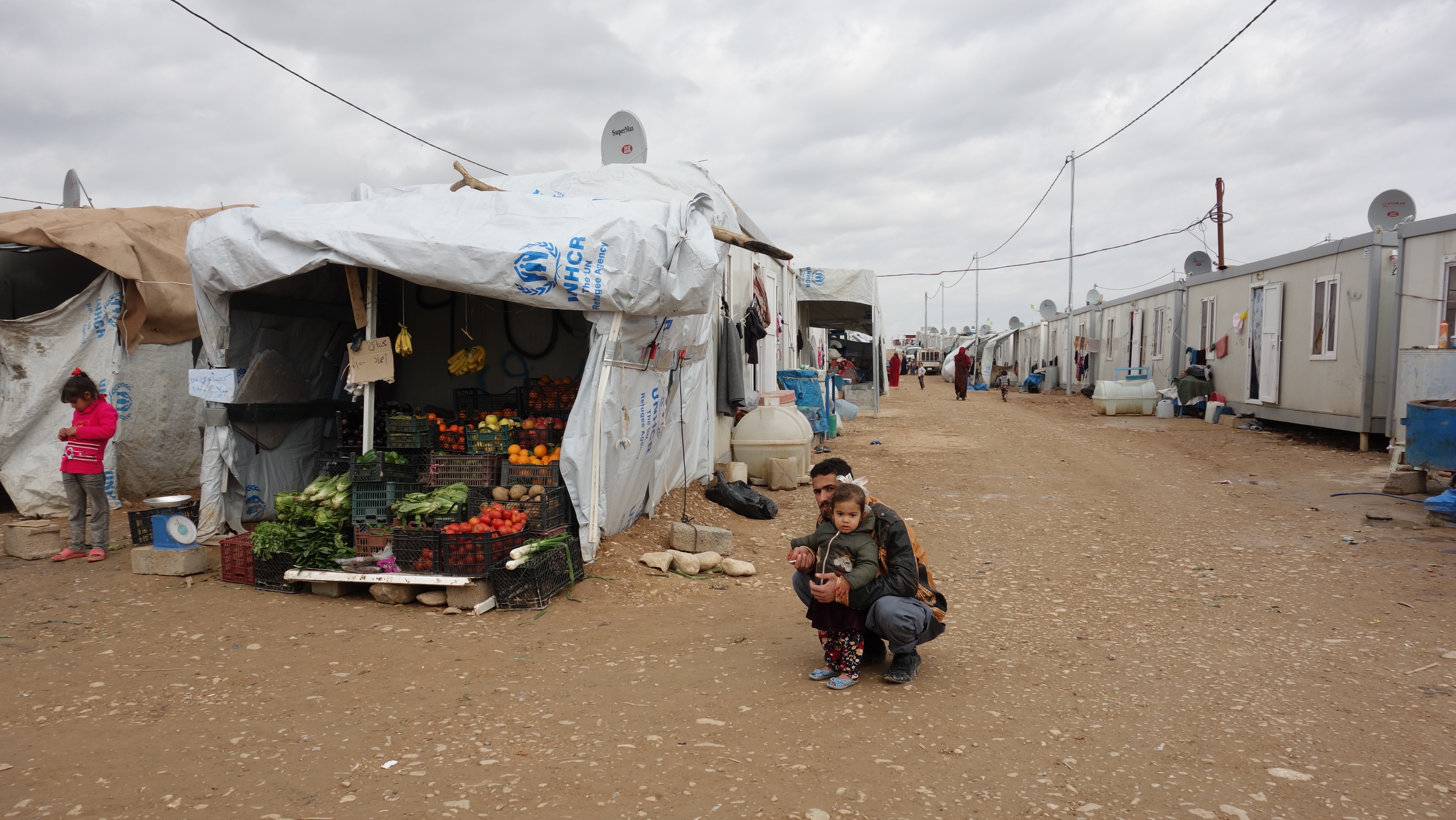 A female child with her dad beside a tent of the UNHCR with fruits and vegetables. 