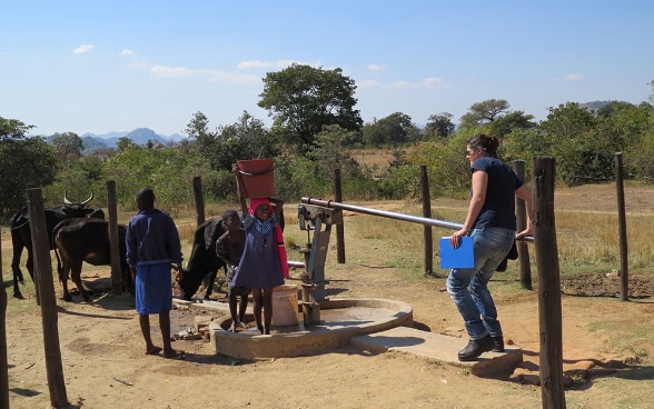Eine Expertin des SKH pumpt Wasser aus einem Brunnen, um den afrikanische Kinder stehen.