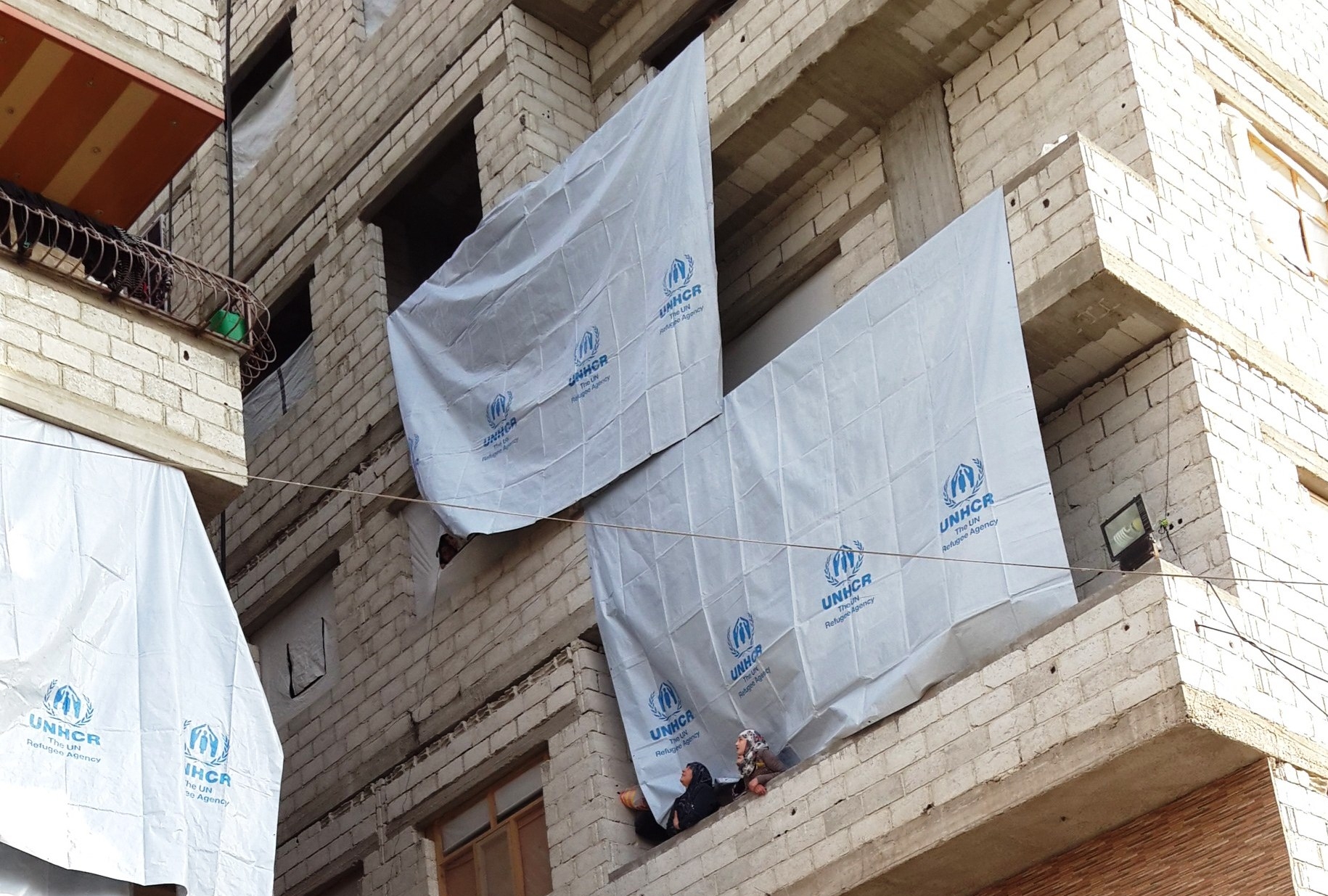 Ein Gebäude mit UNHCR-Fahnen und zwei verschleierten Frauen auf einem Balkon. 