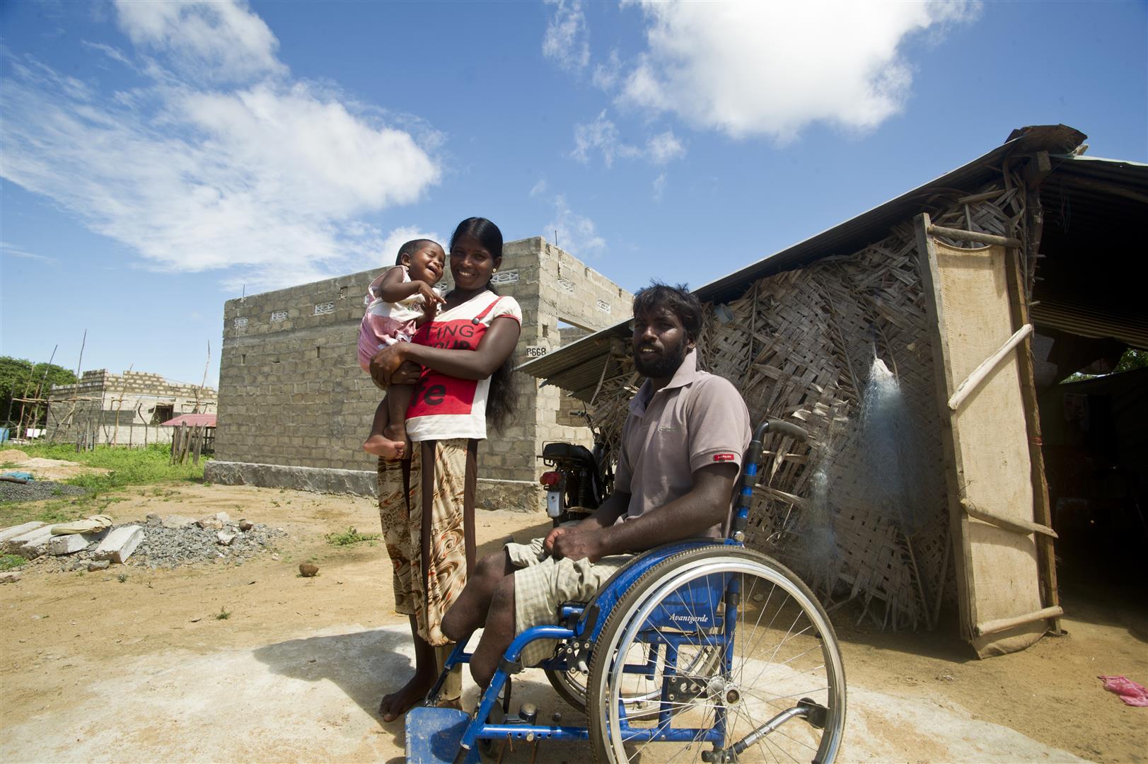 Un uomo in sedia a rotelle, sua moglie e il loro bebè posano davanti alla loro futura casa.