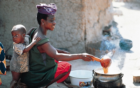 Ein Kind lehnt an eine Frau in Mali, die auf dem Boden das Essen zubereitet