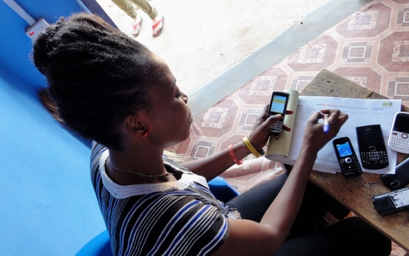 Eine Afrikanerin sitzt an einem Tisch, auf dem mehrere Mobiltelefone liegen, und überträgt Zahlen in eine Tabelle