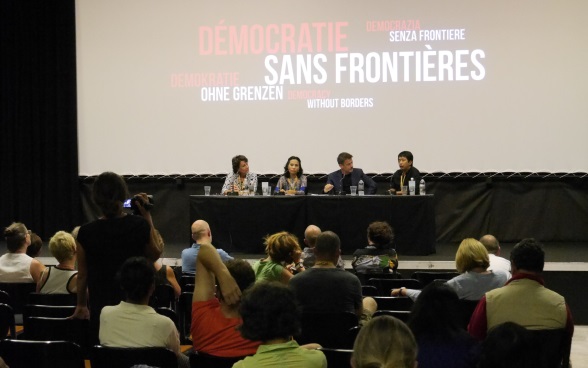 Four people sit at a table in a cinema auditorium. In the foreground, the audience listens to their debate. 