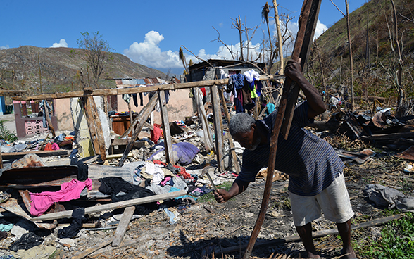 A man holding a hammer stands in front of his destroyed home.