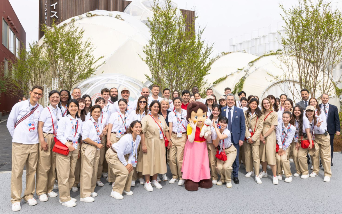 A large group of people in uniform poses with a person dressed as Heidi in front of dome-shaped buildings and trees.
