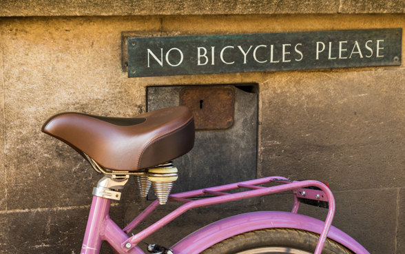 Vélo rose stationné le long d’un mur, sur lequel un écriteau stipule qu’il est interdit de parquer des vélos à cet emplacement.