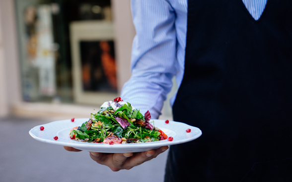 A man in a striped shirt wearing an apron serving a colourful salad. 