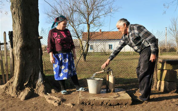 An elderly man and woman filling a bucket with drinking water from a local village tap.