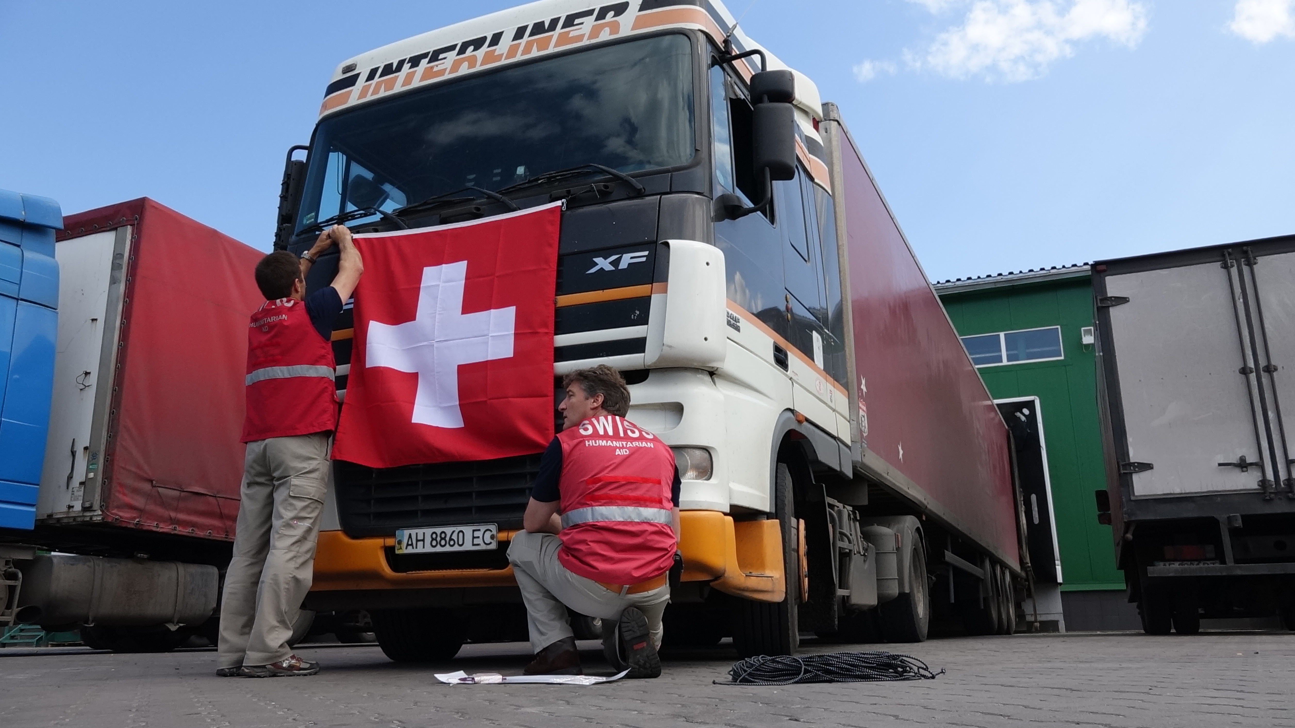 Zwei Mitglieder des Schweizerischen Korps für Humanitäre Hilfe befestigen eine Schweizer Fahne an der Motorhaube eines Lastwagens. 