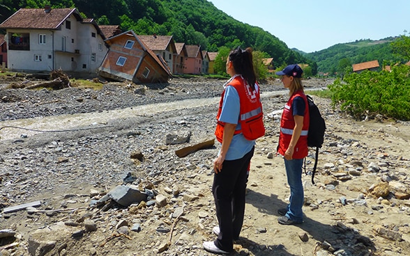 Two women in reflective vests looking at a destroyed house on the banks of a stream.