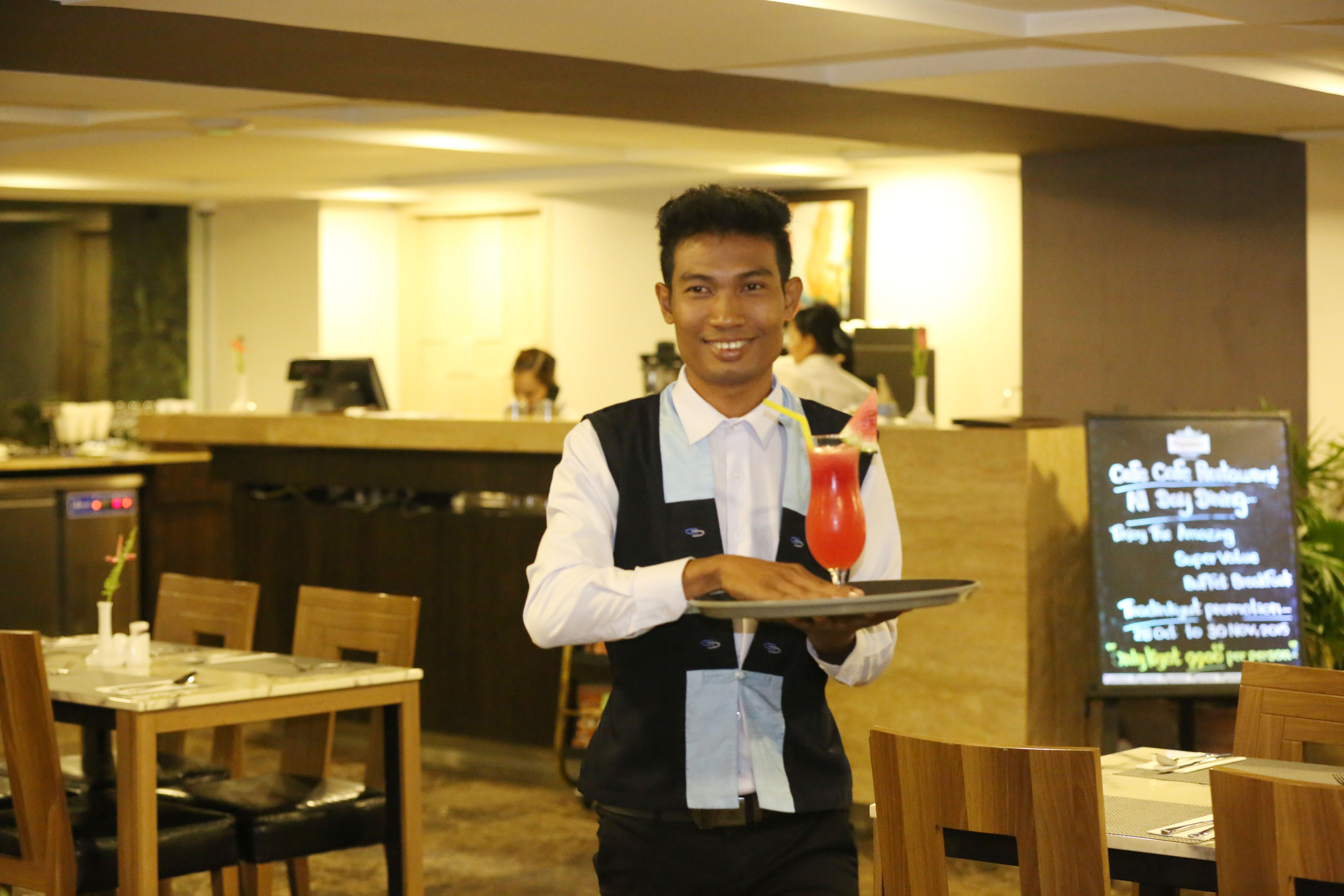 A young waiter smiles as he walks through a restaurant, holding a cocktail on a tray.