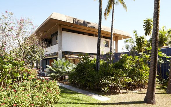 Foreground: bushes and palm trees; background: a two-storey building.