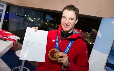 Dario Cologna hält in einer Hand das Glückwunschschreiben von Didier Burkhalter und in der anderen seine Goldmedaille.