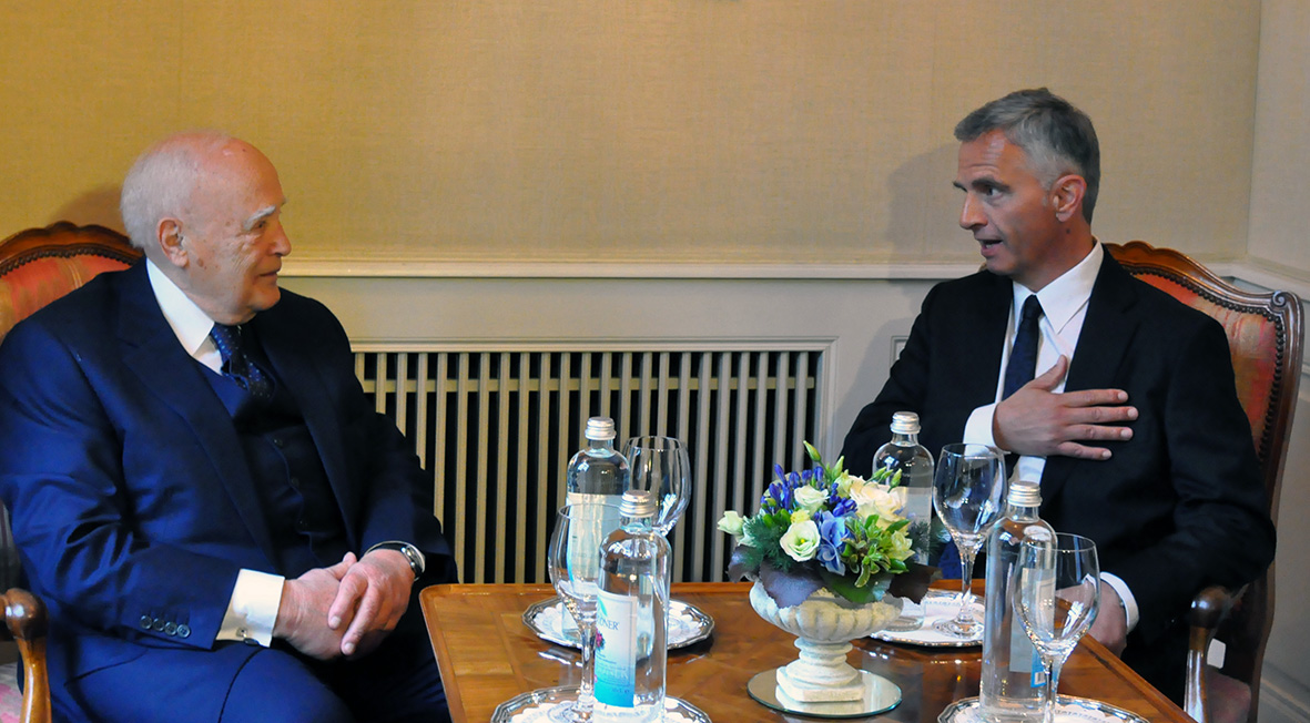 Didier Burkhalter and Karolos Papoulias sitting at a table together talking. 
