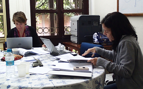 Two employees of the Swiss embassy to Nepal working at a table strewn with laptops and files.
