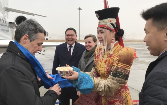 Eine Frau in einem traditionellen Kleid überreicht Bundesrat Ignazio Cassis beim Verlassen des Flugzeugs eine Schale.