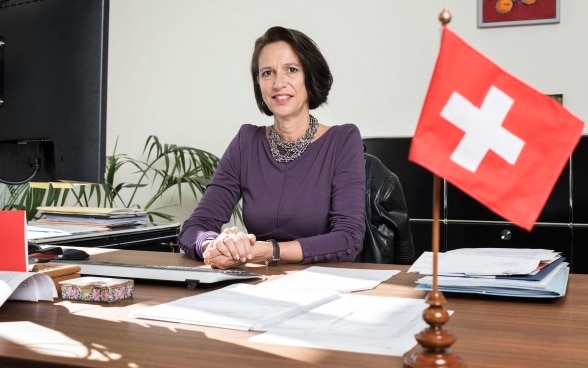 Eine Frau mit schwarzem Haar, Christine Schraner Burgener, sitzt hinter einem Schreibtisch, auf welchem eine kleine Fahne der Schweiz steht.