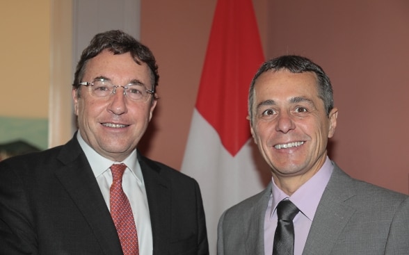 Ignazio Cassis und Achim Steiner stehend vor der Schweizer Fahne.