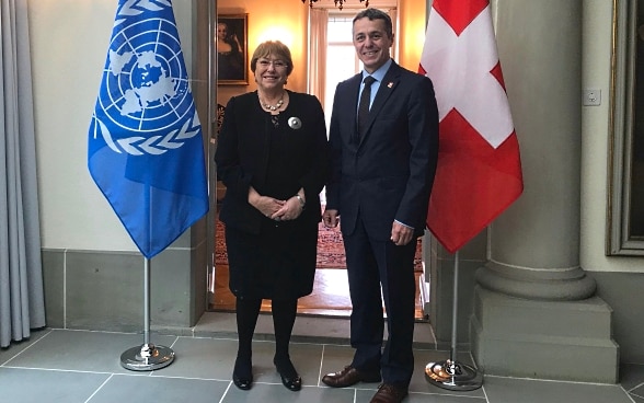 Bundesrat Ignazio Cassis und die UNO-Hochkommissarin für Menschenrechte Michelle Bachelet posieren für ein Foto. Im Hintergrund sind die Flaggen der Schweiz und der UNO zu sehen.