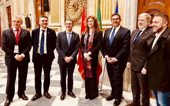 Federal Councillor Ignazio Cassis (third from left), a woman and five other men stand in a semicircle in front of the three flags.