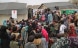 Several women, men and children are gathered in front of two tents with a stage.