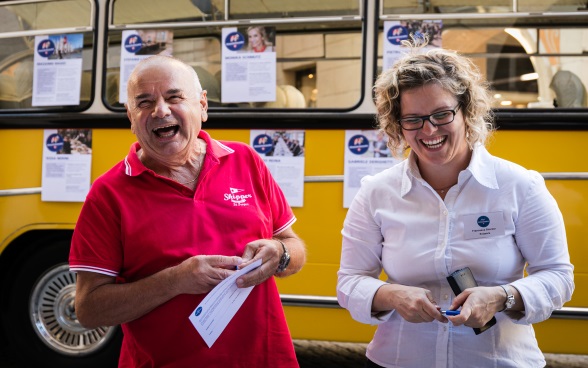 Un visiteur en chemise rouge rit avec une ambassadrice; à l'arrière-plan se trouve le bus postal Meet the Ambassador