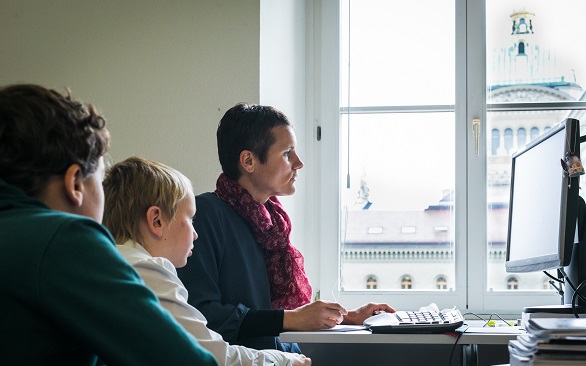 Zwei Kinder schauen einer Frau bei der Arbeit an einem Computer über die Schulter.