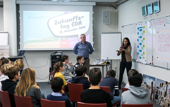 Des enfants écoutent une femme présenter le travail de l'Aide humanitaire de la Suisse.