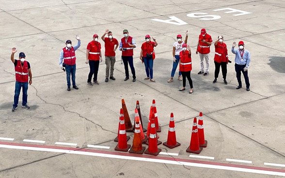 Blick aus dem Flugzeug auf eine Gruppe von Botschaftsmitarbeitern in roten Jacken, die auf dem Flugfeld steht und winkt. 