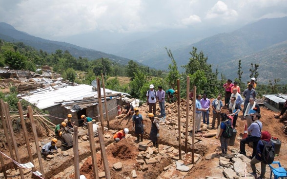 Dans un paysage boisé et vallonné, des ouvriers népalais reconstruisent les murs d’une maison. 