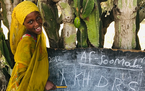  A little girl with a textbook sits at a wooden desk in a classroom and looks at the camera. 