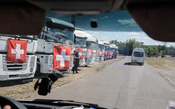 Zehn Lastwagen mit Schweizer Flaggen stehen aufgereiht auf einem Feld in der Ukraine. 