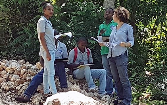 Constance Jaillet stands under trees with papers in her hand and talks to local people.