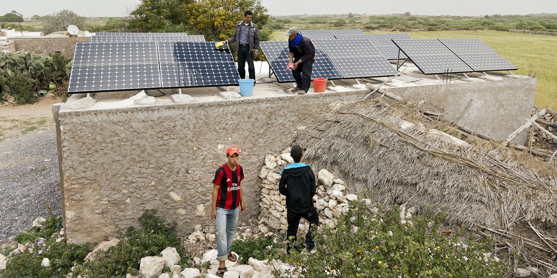 Deux hommes se tiennent sur un bâtiment en pierre et lavent des panneaux solaires. A côté de la maison, il y a deux autres hommes.