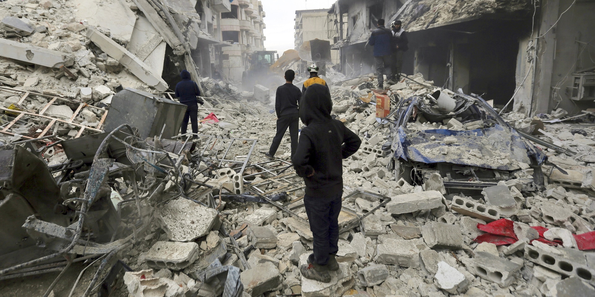 Des civils et un casque blanc parcourent un amas de décombres dans une rue de la ville d’Idlib. Au premier plan, un jeune garçon encapuchonné de dos regarde au loin.