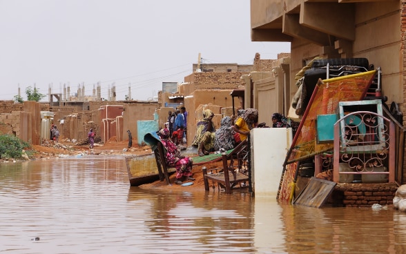 Some people stop at the side of the road submerged by water. Next to them some recovered furniture.