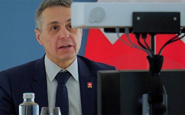 Federal Councillor Ignazio Cassis gives a speech in front of a camera during a virtual conference. Behind him, the Swiss flag.