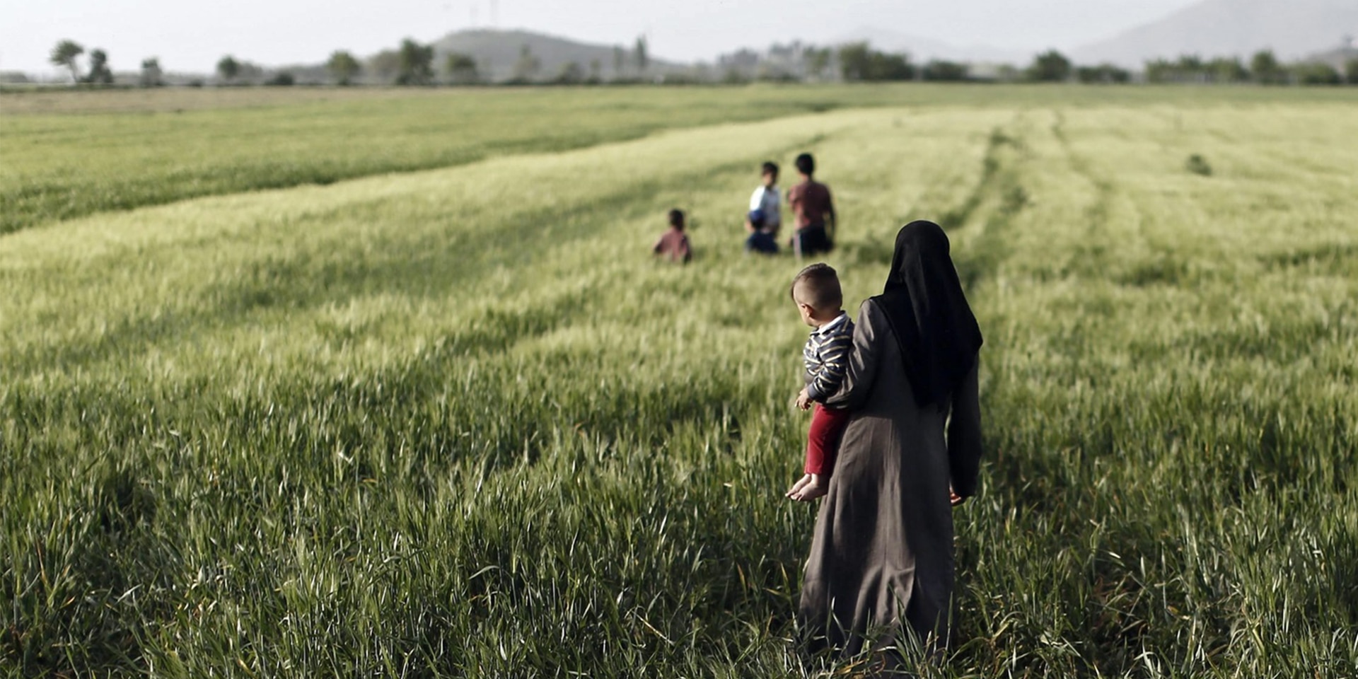 Eine Frau steht mit einem Kleinkind auf dem Arm in einem Feld mit hohem Gras und schaut Kindern in der Ferne beim Spielen zu.