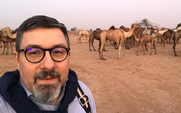  Il volto di un uomo in primo piano e una mandria di cammelli sullo sfondo.