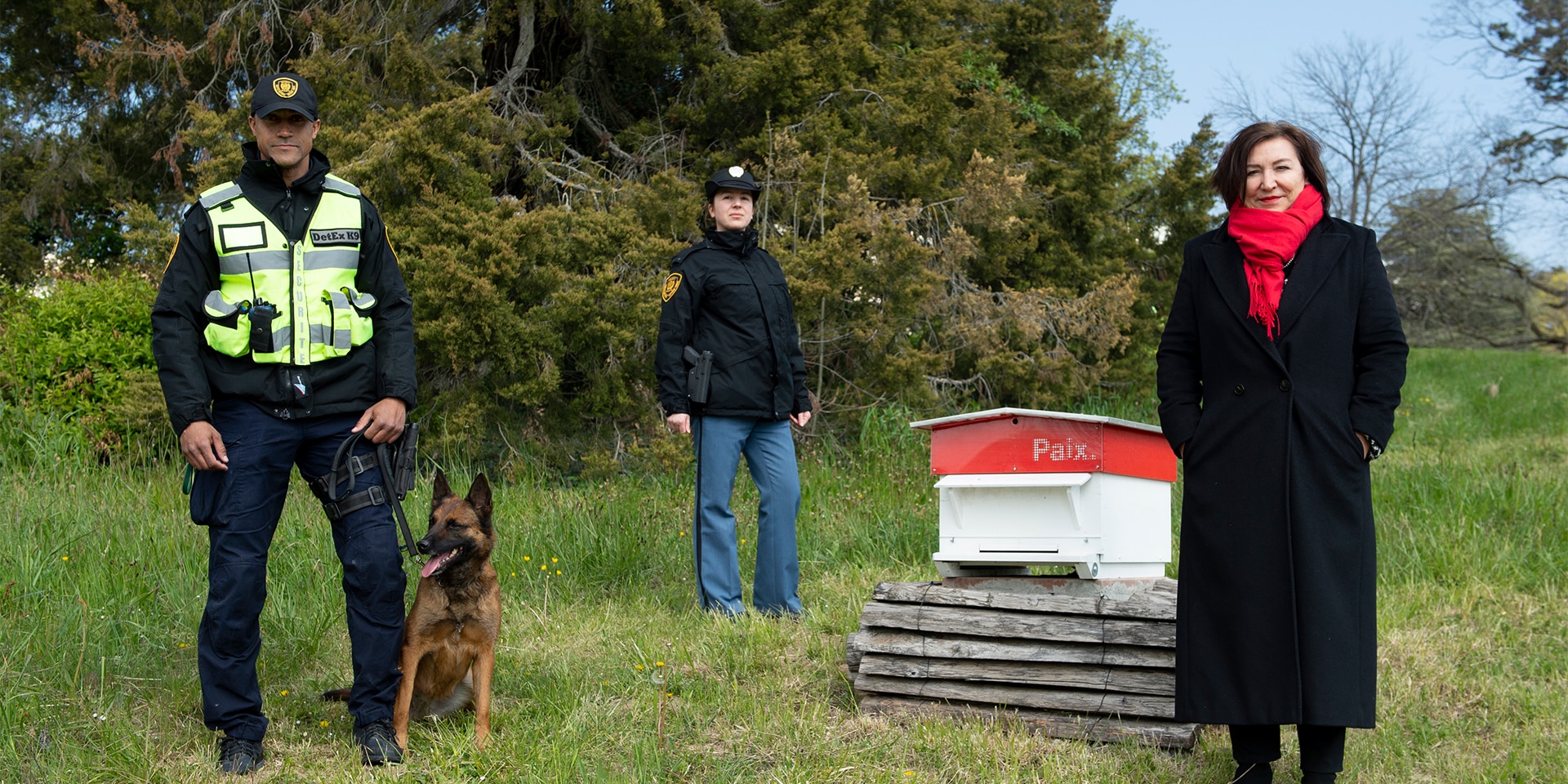 Auf dem Rasen des Gebäudes der Vereinten Nationen steht Anne-Lise Favre Pilet neben zwei UN-Agenten und einem Bombenspürhund. 