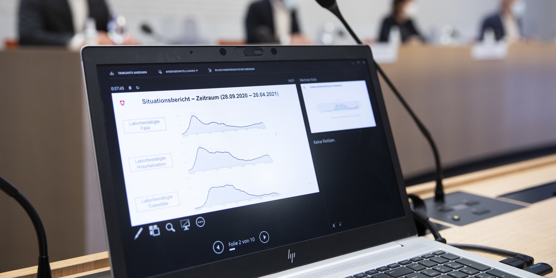 Trend curves displayed on a computer screen with the members of the Federal Council in the background in the Federal Palace Media Centre.