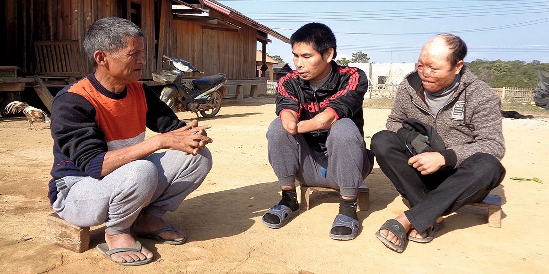 Three Lao cluster munition victims sit on the ground in front of a farmhouse.
