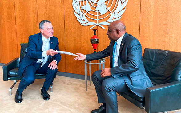 Sullo sfondo dell'emblema dell'ONU, il consigliere federale Ignazio Cassis consegna un documento al presidente dell'Assemblea generale dell'ONU Abdulla Shahid (entrambi seduti).
