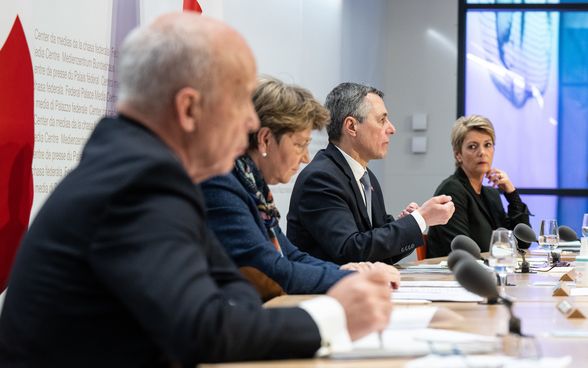 Four members of the Federal Council sit at a large podium. Media representatives are seated opposite them. 