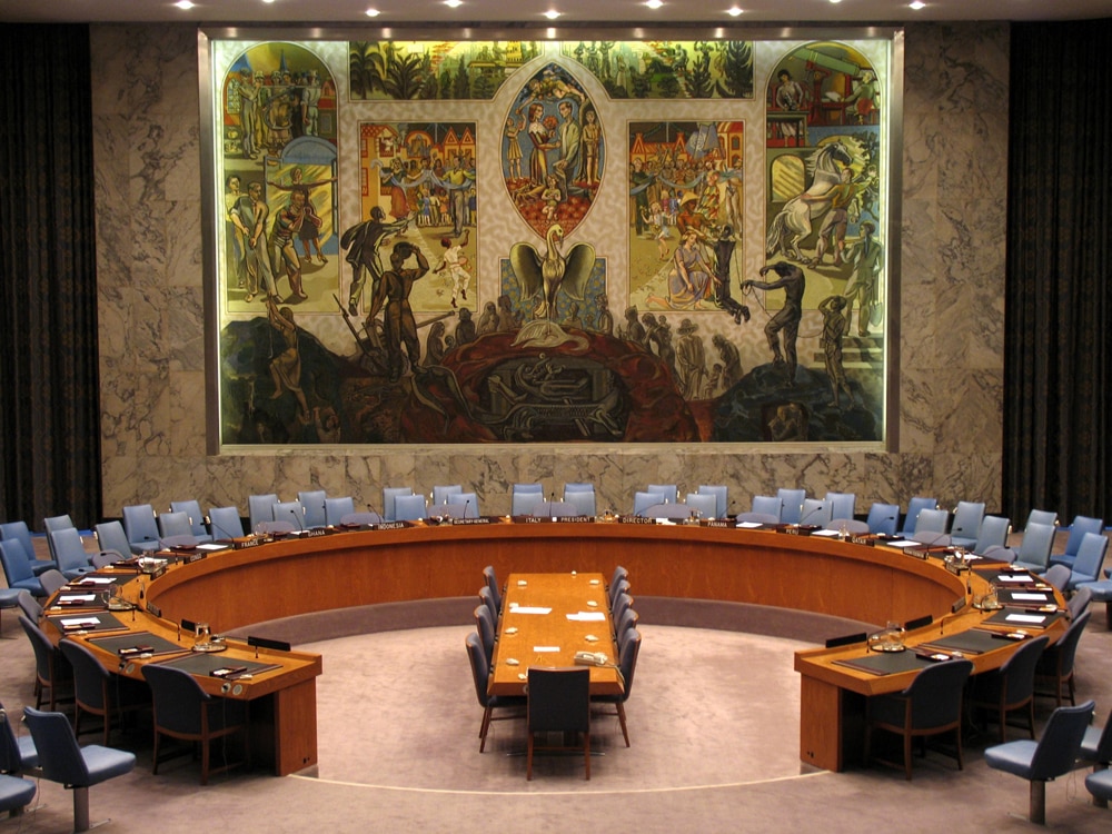  Une table en bois en forme de fer à cheval entourée de nombreuses chaises. Une peinture murale figure à l’arrière-plan. Elle représente un phénix symbolisant un nouveau départ après la Seconde Guerre mondiale.