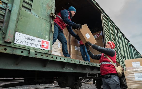  An der offenen Tür eines Güterwagens stehen Männer in roten Jacken und laden Schachteln ein.