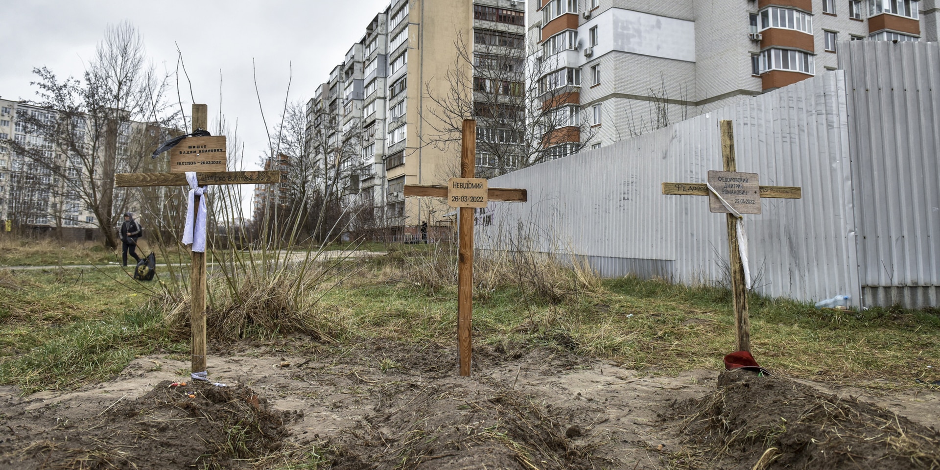 Three graves with wooden crosses and names can be seen in the foreground. Large houses are in the background.