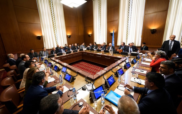 A group of men and women facing each other at a large wooden table with computer monitors.