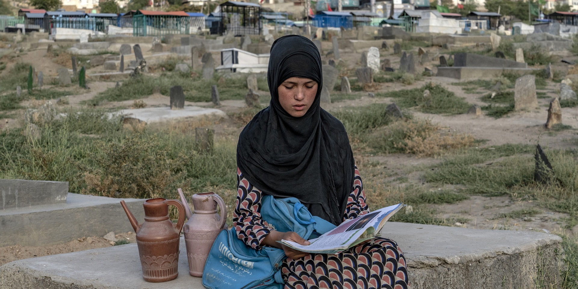 Une fillette assise sur une dalle en pierre lit un livre. Elle a près d’elle un sac estampillé ONU Femmes, et on aperçoit en arrière-plan des habitations sommaires.