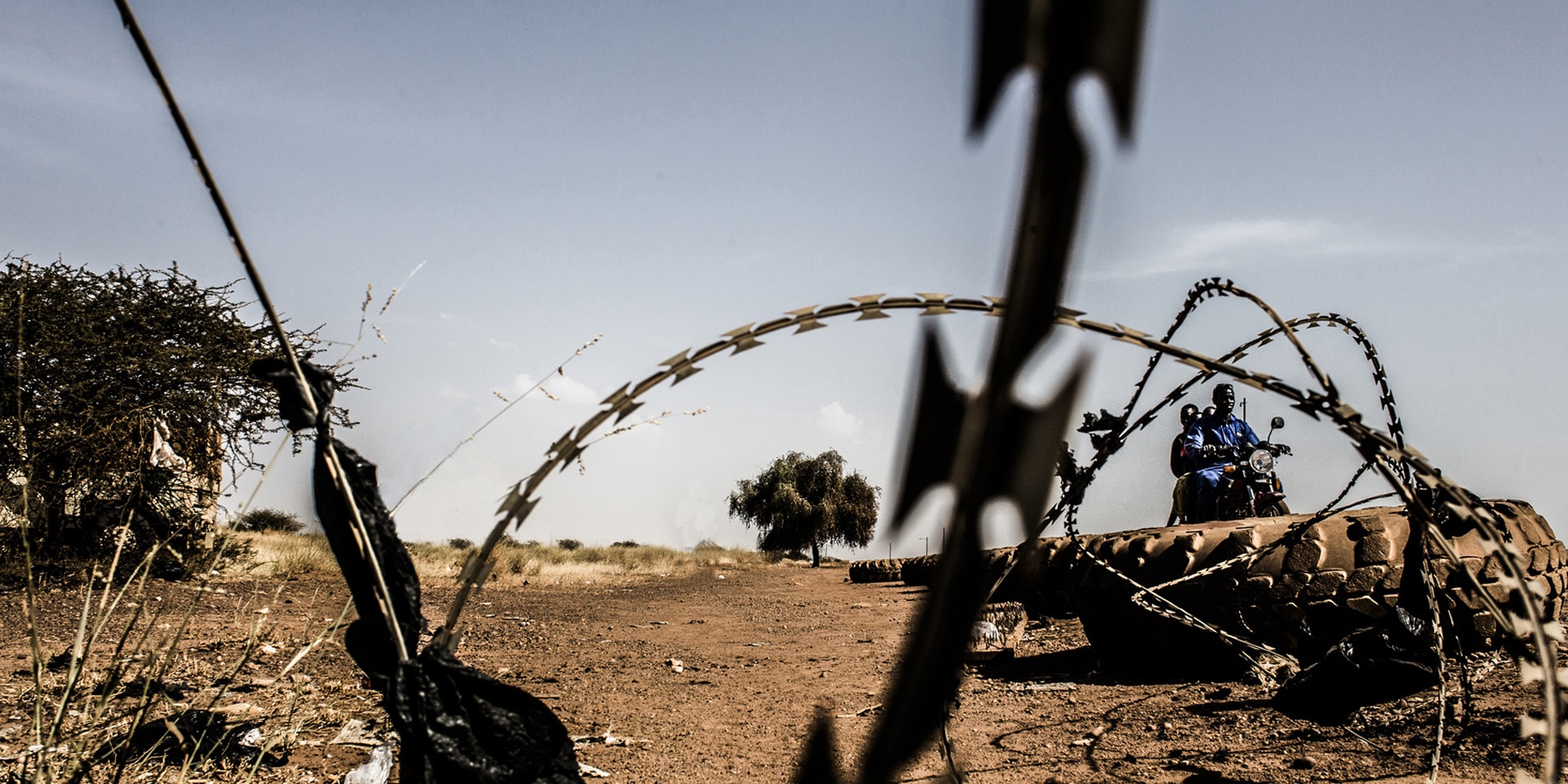 Un fil de fer barbelé et des pneus sur une route sablonneuse en Afrique. En arrière-plan, deux personnes à moto.